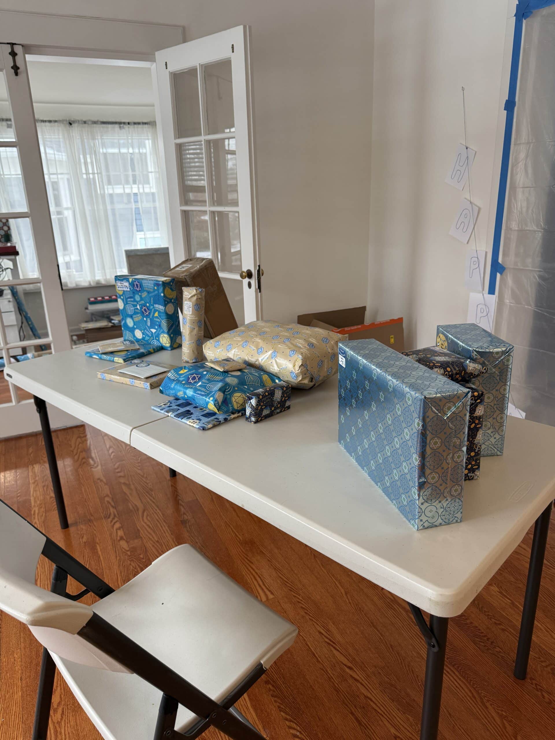 A folding table holds several wrapped gifts in blue and gold paper, with two white chairs nearby. A banner with the word “HAPPY” hangs on the wall in a bright, sunlit room with wooden floors.
