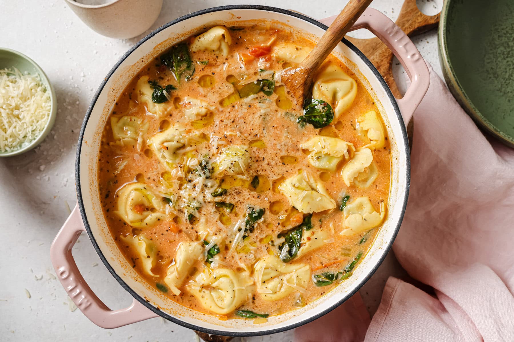 A creamy tortellini soup with spinach and tomatoes simmers in a pink Dutch oven. A wooden spoon rests in the soup, and grated cheese is sprinkled on top. A small bowl of cheese and a pink napkin are nearby.