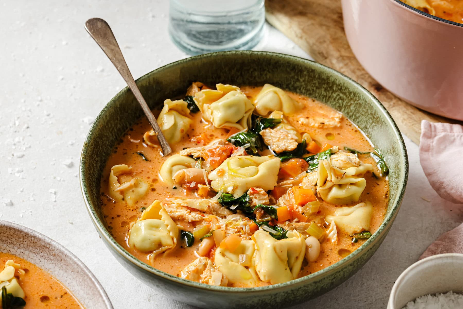 A green bowl filled with creamy tomato-based tortellini soup, featuring spinach, pieces of chicken, carrots, and tortellini pasta, with a spoon resting in the bowl. A pot and a glass of water are nearby.