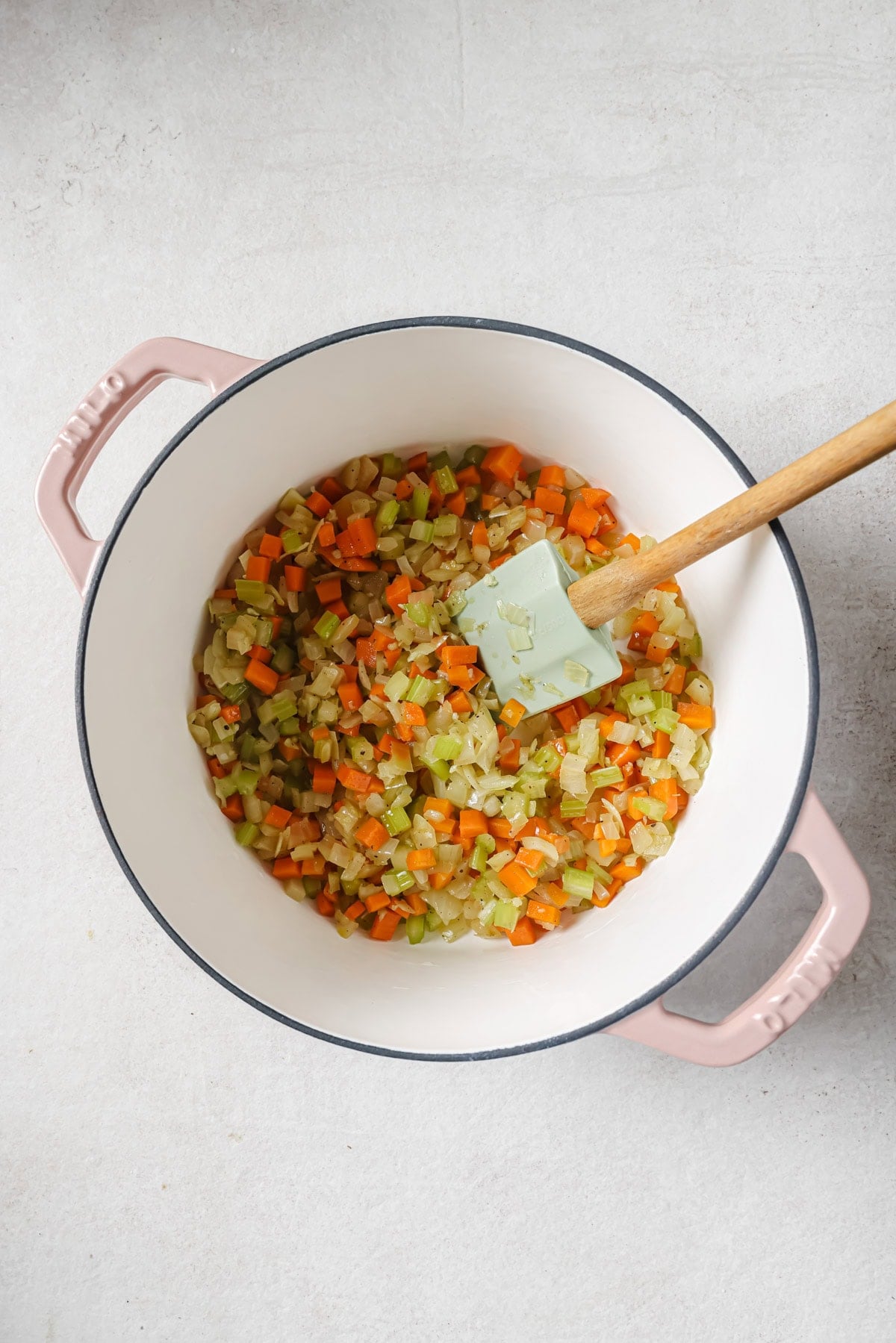 A white pot with pink handles contains sautéed chopped onions, celery, and carrots. A light blue spatula with a wooden handle rests inside the pot on a light-colored surface.