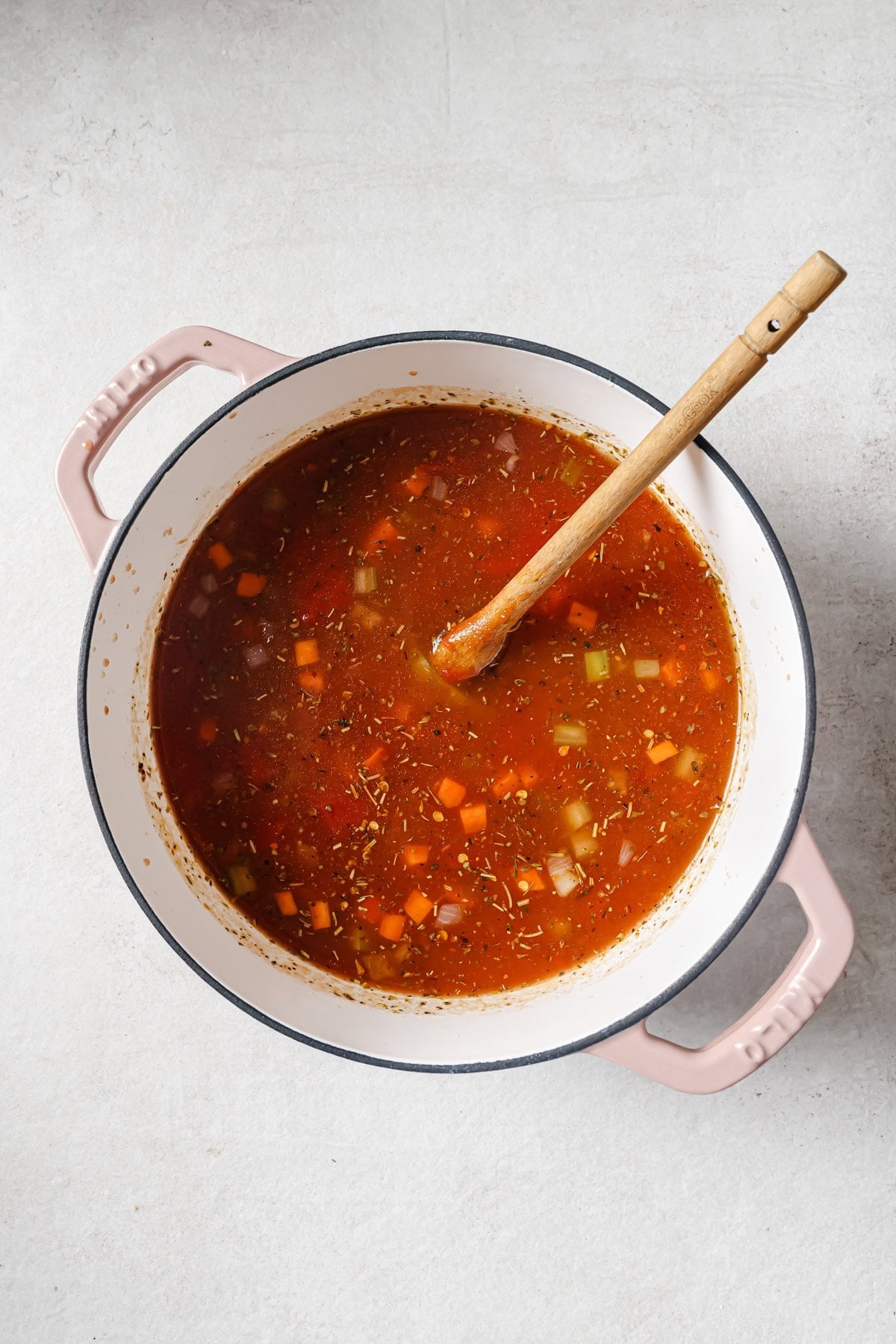 A large white pot with pink handles filled with vegetable soup, containing visible chunks of carrot, celery, and other vegetables in a red broth, with a wooden spoon resting inside.