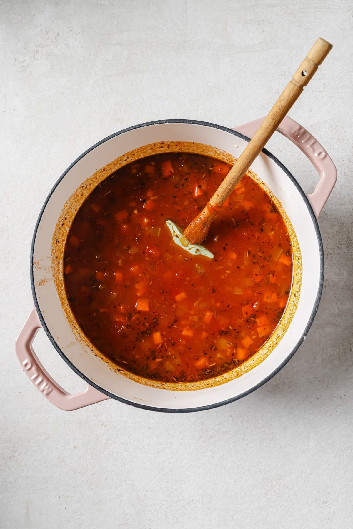 A pot of tomato-based soup with diced vegetables and herbs, stirred with a wooden spoon, sits on a light-colored surface.