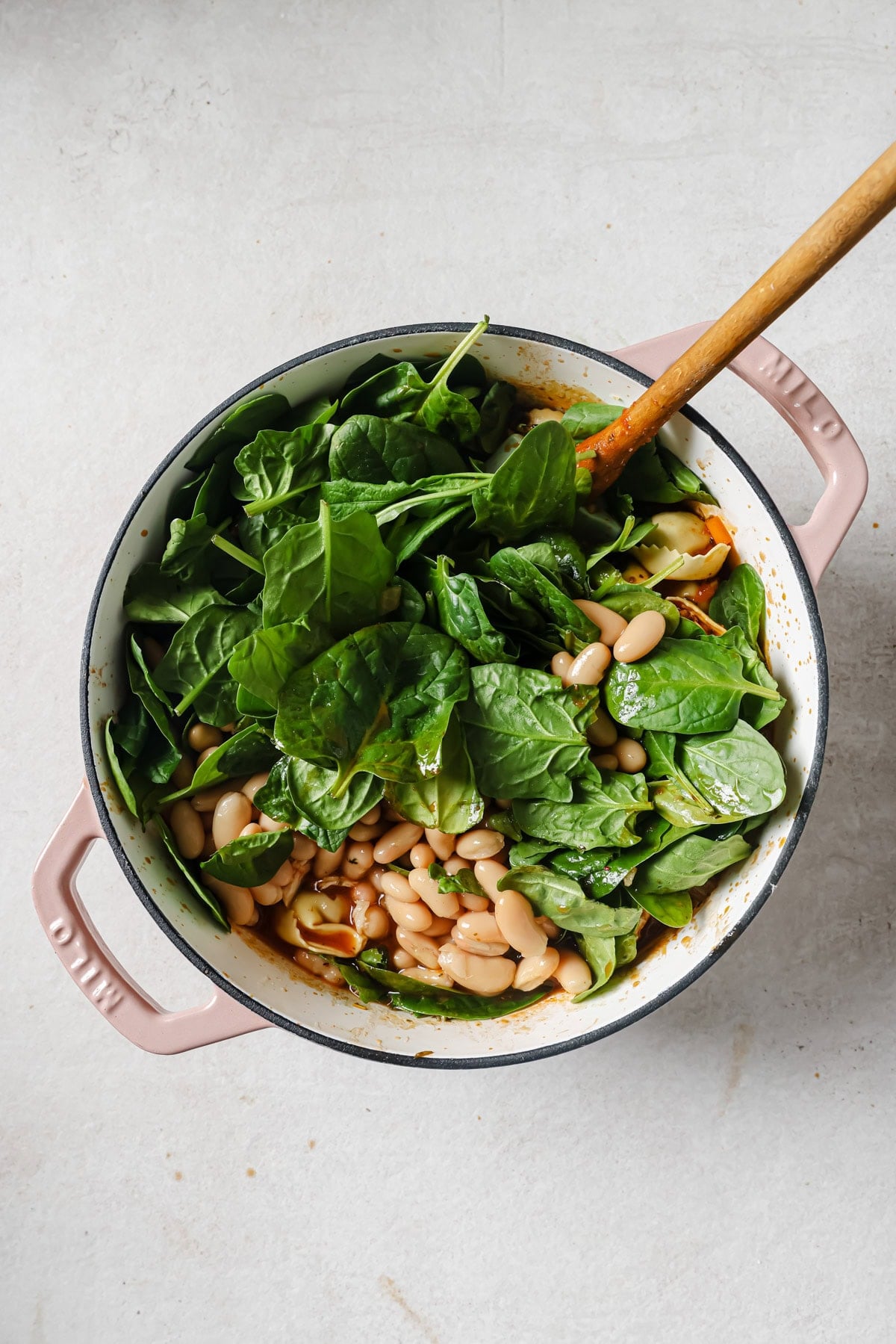 A pink-handled pot filled with fresh spinach leaves, white beans, and other ingredients being mixed with a wooden spoon on a light gray surface.