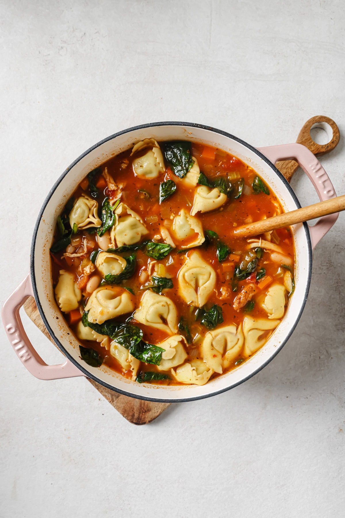 A pot of tortellini soup with spinach and beans in a tomato-based broth sits on a wooden board, stirred with a wooden spoon.