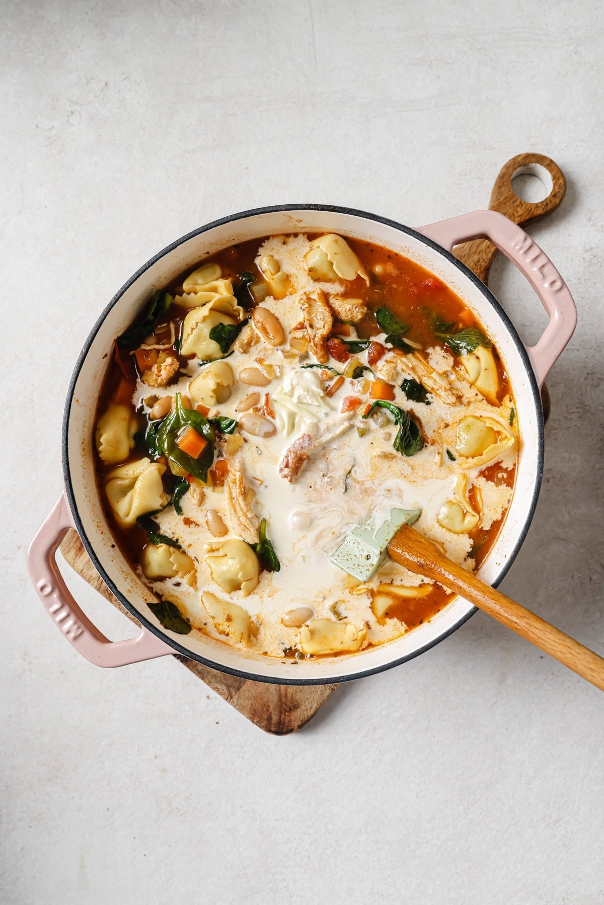 A pot of creamy tortellini soup with spinach and chunks of vegetables is sitting on a wooden trivet. A spatula rests in the pot, and cream has just been poured in, swirling into the soup.
