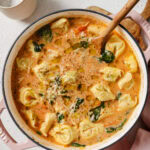 A creamy tomato-based tortellini soup with spinach, grated cheese, and herbs is simmering in a large pot with a wooden spoon, surrounded by plates, a bowl of grated cheese, and a glass of water.