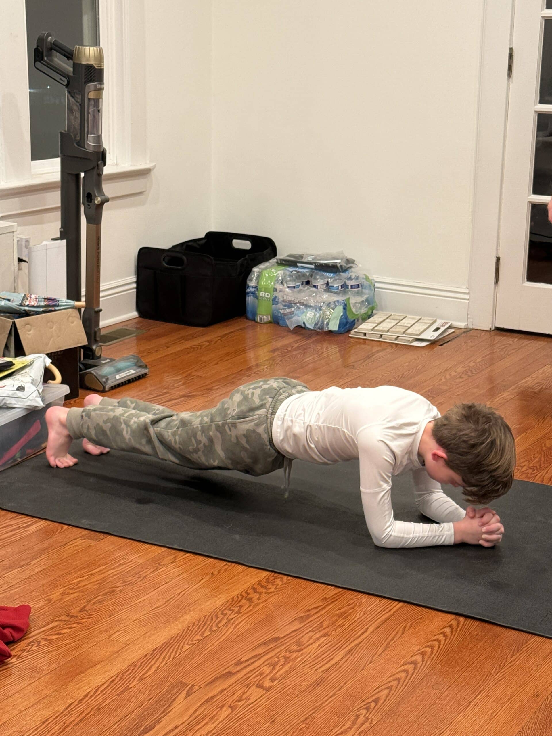 A young boy in a white shirt and camouflage pants holds a plank position on a black exercise mat in a room with hardwood floors, near stacked water bottles and various household items.