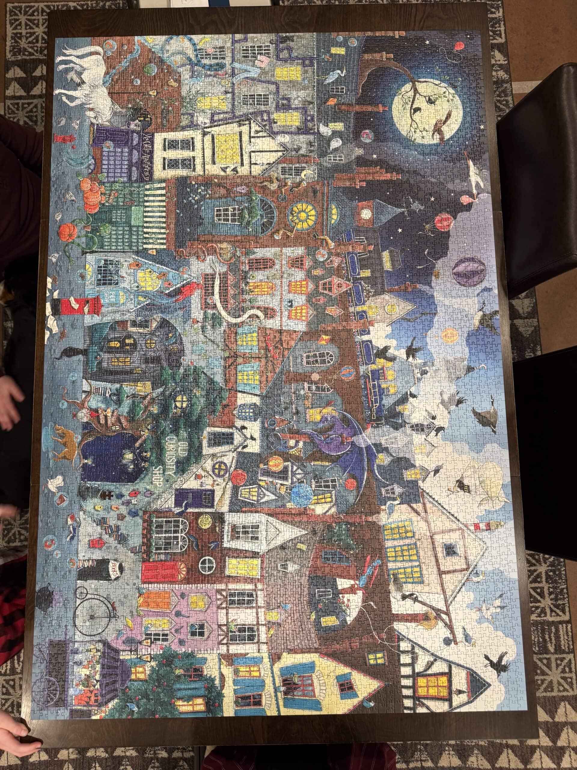 A colorful, completed jigsaw puzzle on a table shows a whimsical town scene at night, with houses, people, animals, a large moon, and various magical and fantasy elements. Two peoples hands are visible at the edges.