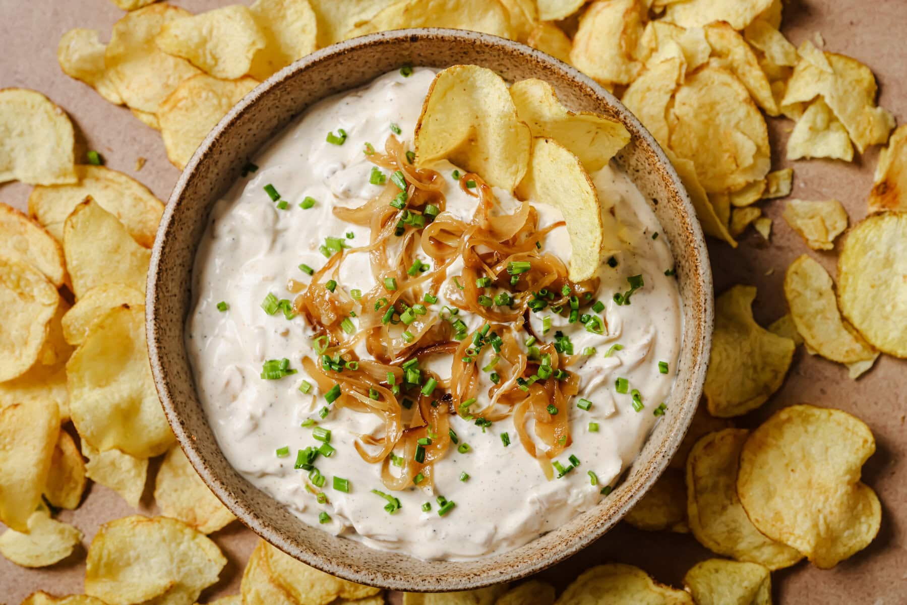 A bowl of creamy onion dip topped with caramelized onions and chopped chives, surrounded by scattered potato chips on a light surface.