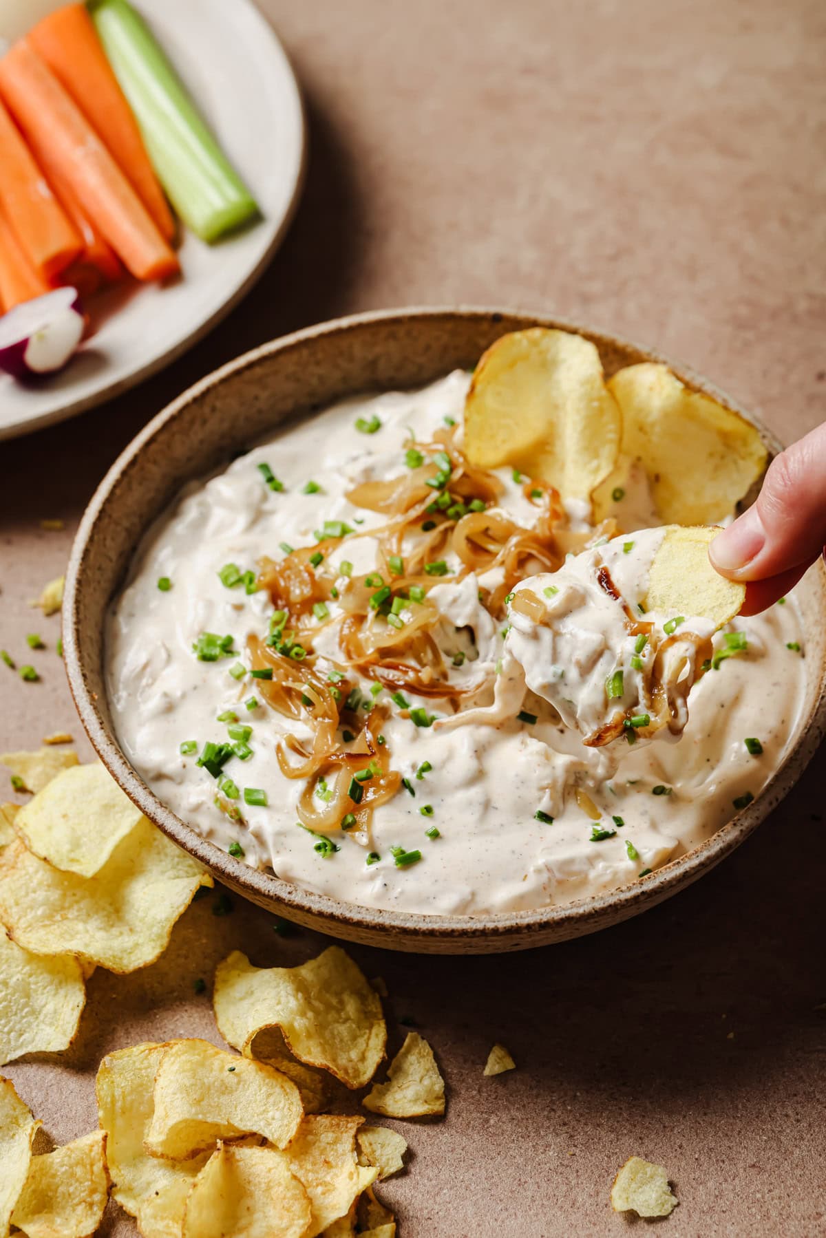 A hand dips a potato chip into a bowl of creamy onion dip topped with caramelized onions and chopped chives; veggie sticks and more chips are nearby.