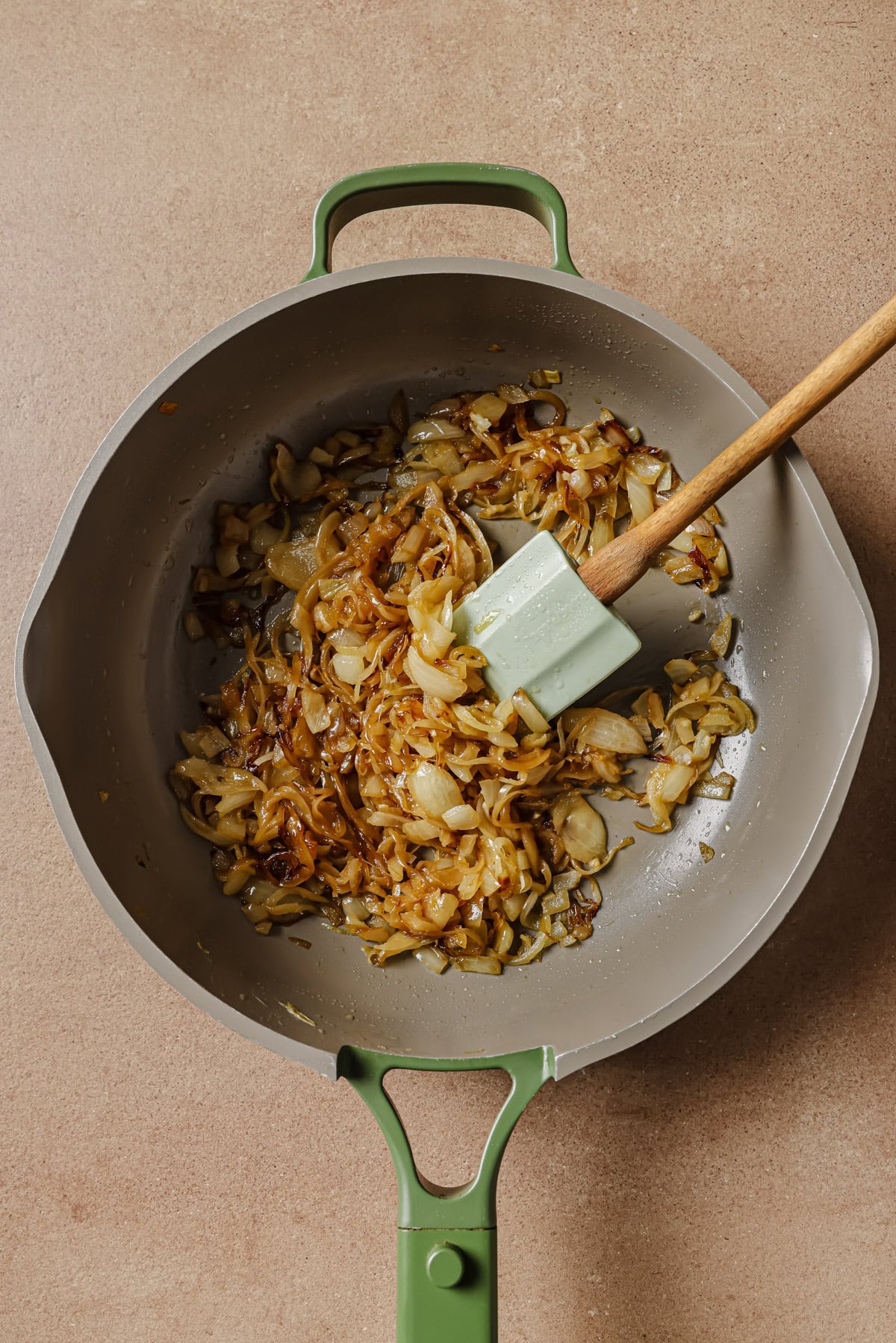 A green-handled pan containing caramelized onions being stirred with a green spatula, set on a light brown countertop.