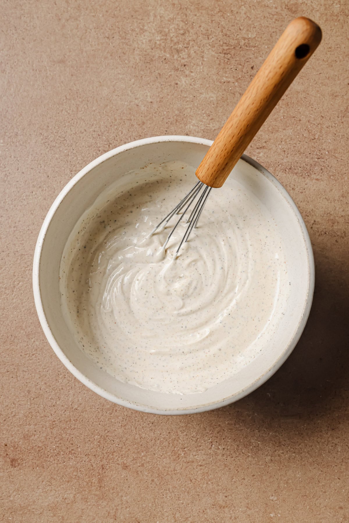 A whisk with a wooden handle rests in a white bowl filled with creamy, light-colored sauce or batter on a brown countertop.