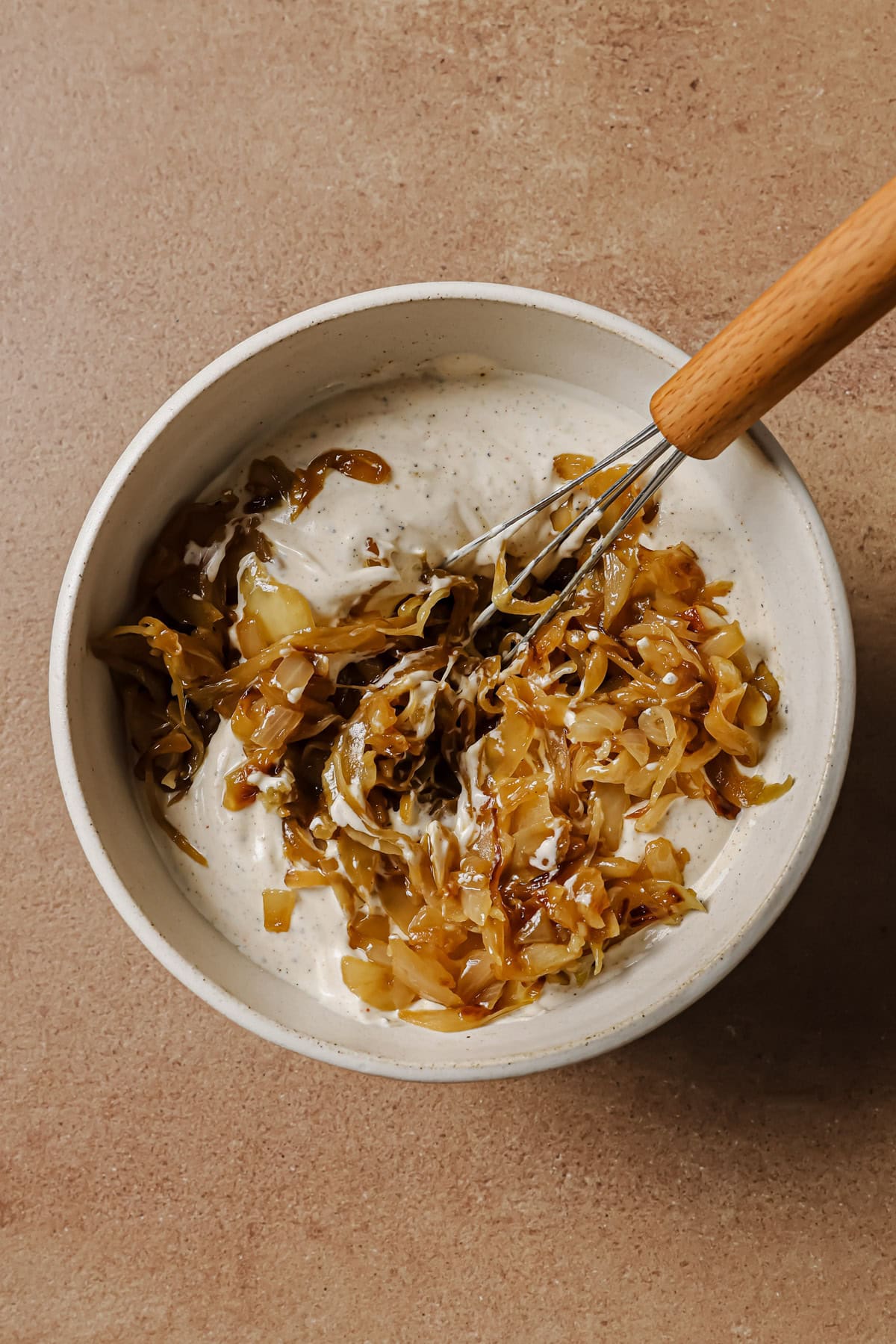 A white bowl with creamy dip being mixed with caramelized onions using a small whisk, set on a light brown surface.