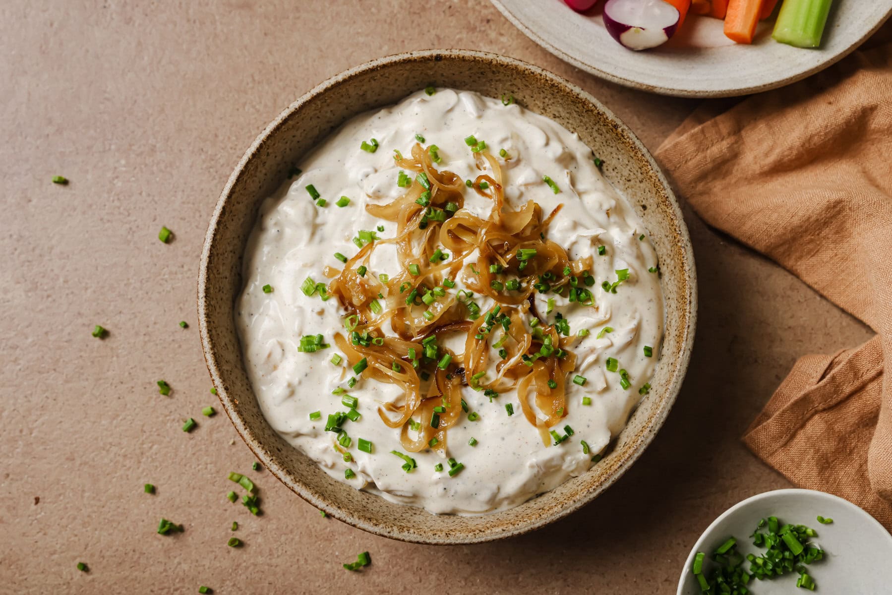 A bowl of creamy dip topped with caramelized onions and chopped chives sits on a light brown surface, surrounded by a cloth napkin, a partial plate of vegetable sticks, and a small dish of extra chives.