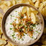 A bowl of creamy dip topped with caramelized onions and chopped chives, surrounded by crispy potato chips, with a few chips dipped into the bowl.
