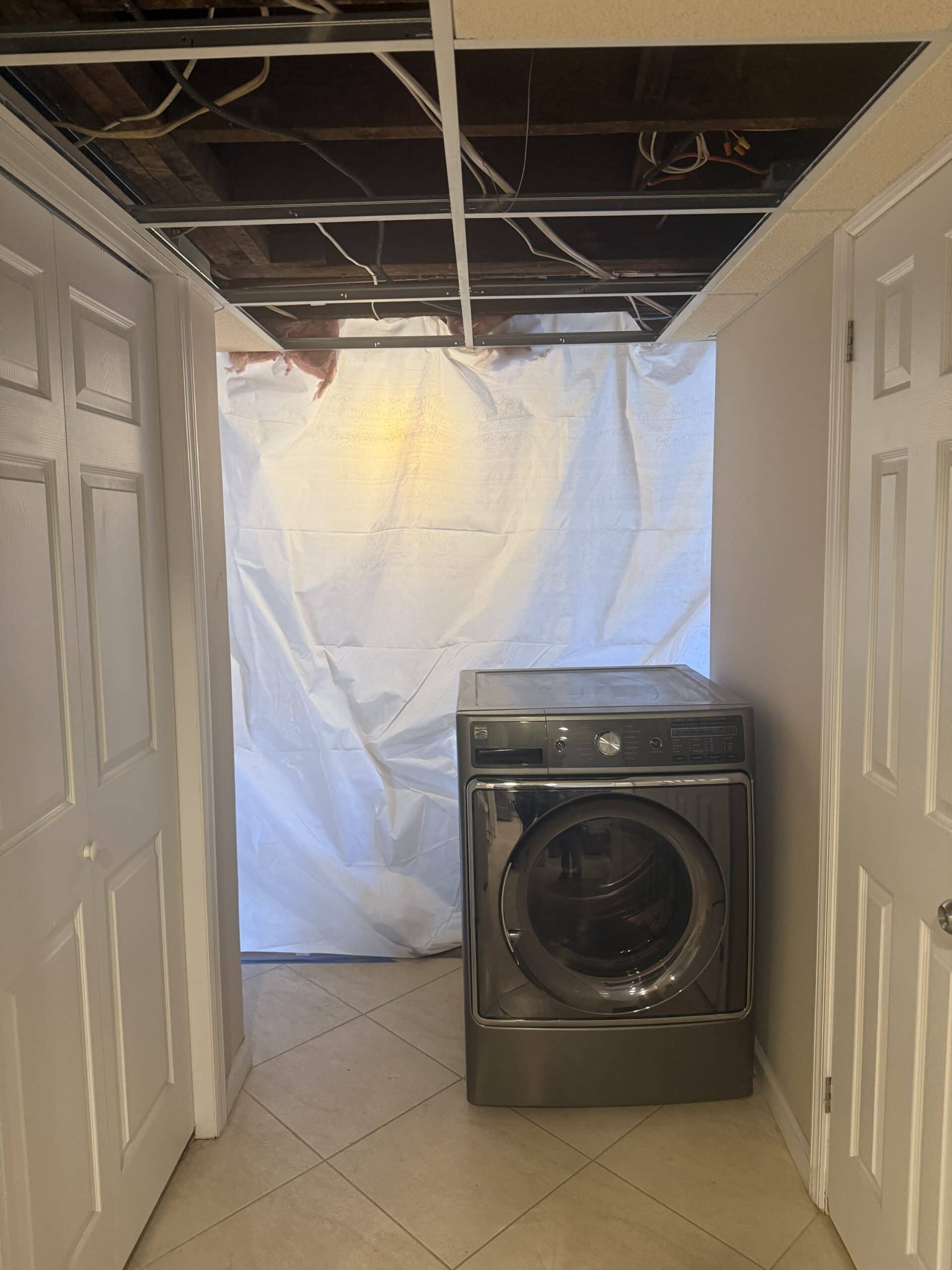 A laundry area with a front-loading washing machine, beige tiled floor, and white walls. The ceiling above is partially missing, exposing pipes and wires, and a white plastic sheet covers the back wall.