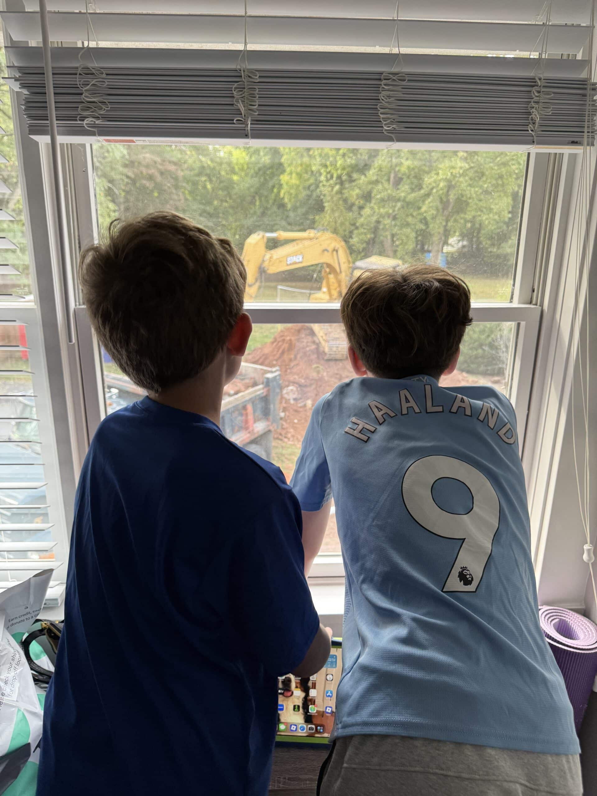 Two boys look out a window at a yellow excavator working outside. One wears a sports jersey with HAALAND 9 on the back, and both appear fascinated by the construction scene through the glass.