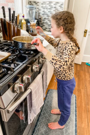 A young girl with braided hair, wearing a leopard print shirt and blue sweatpants, stands barefoot on a kitchen mat, stirring food in a pot amid kitchen utensils—proof of living through construction with kids.