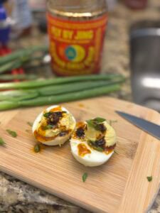 A halved miso-sesame deviled egg topped with chili oil and chopped green onions sits on a wooden cutting board next to a knife, fresh green onions, and a jar of chili oil in the background.