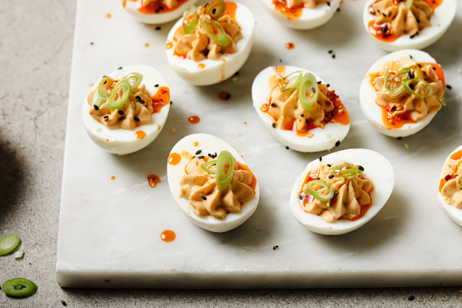 Deviled eggs arranged on a white marble slab, topped with creamy filling, sliced green onions, sesame seeds, and a drizzle of orange sauce. Some sauce and seeds are scattered around the eggs.