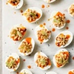 Halved deviled eggs arranged on a marble surface, topped with a creamy filling, sliced green onions, black sesame seeds, and a drizzle of orange sauce.