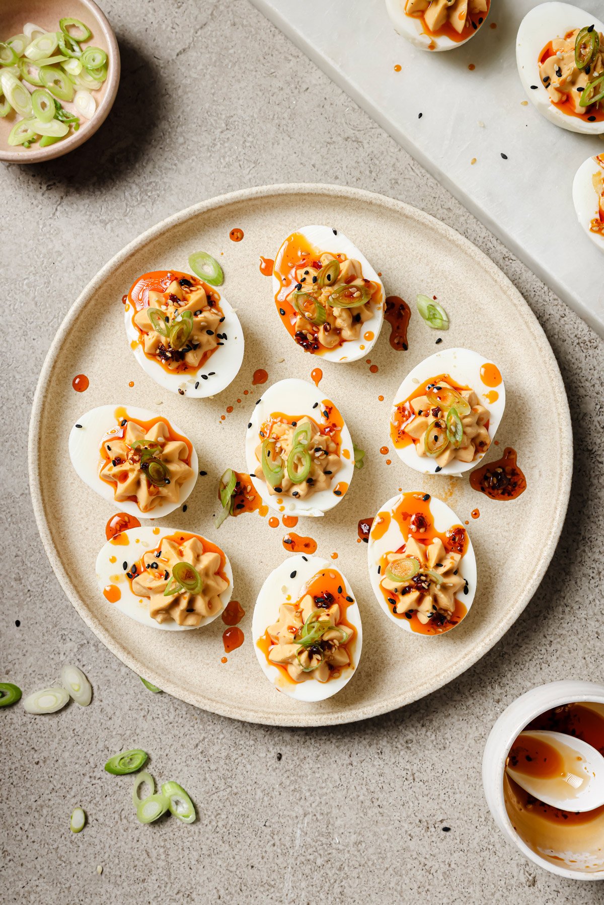 A plate of deviled eggs topped with sliced green onions, black sesame seeds, and a drizzle of chili oil, with extra toppings and ingredients scattered around on a light surface.