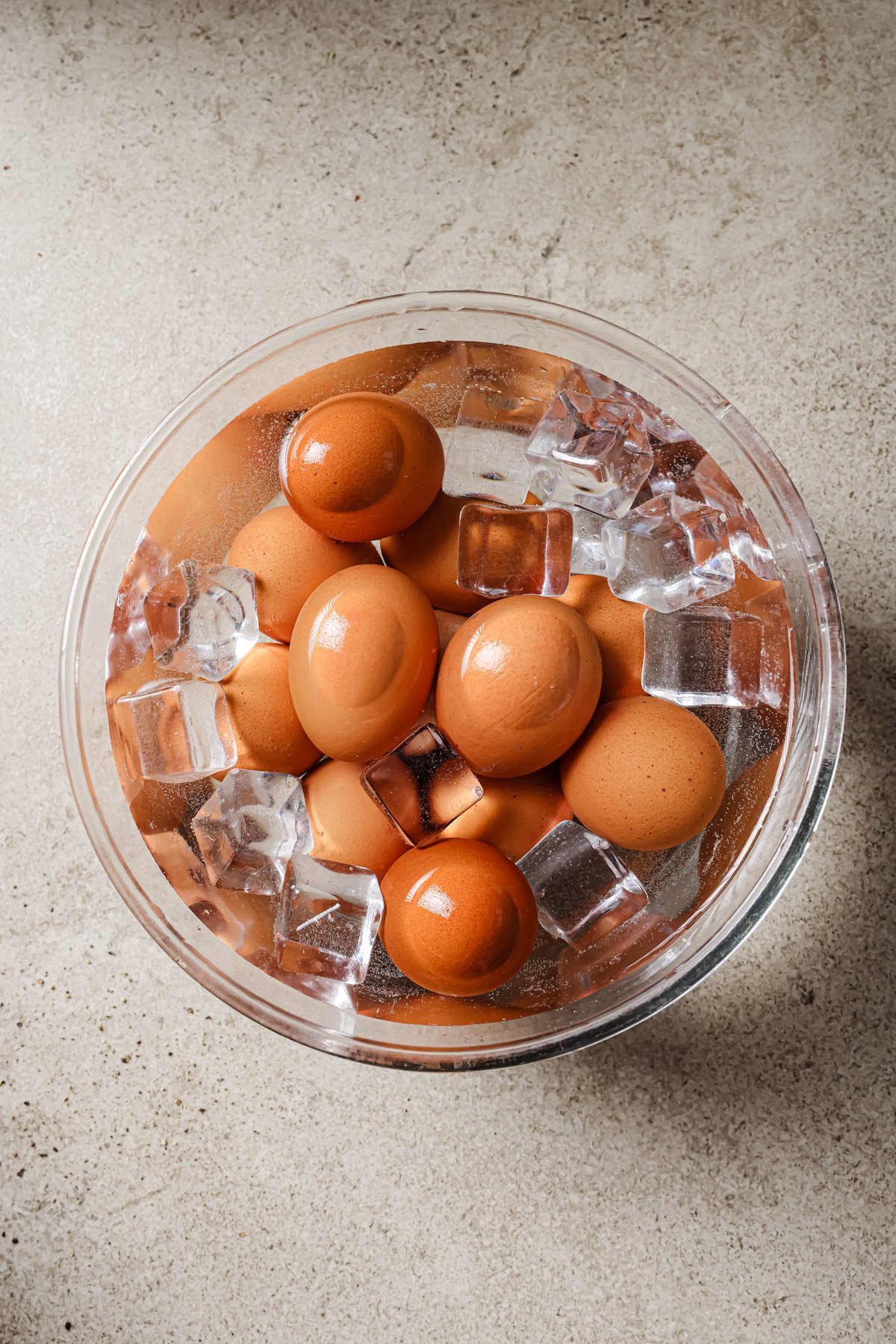 A clear bowl filled with brown eggs and ice cubes sits on a light-colored countertop, showing the eggs being chilled in an ice bath.