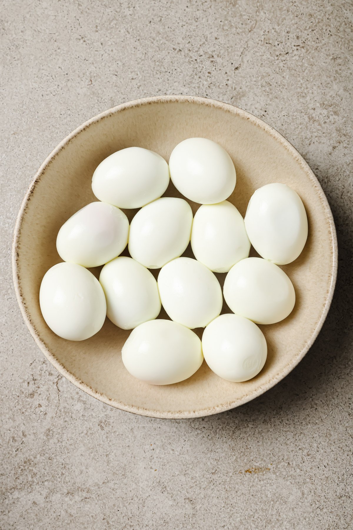 A beige bowl filled with twelve peeled hard-boiled eggs sits on a light gray stone surface.