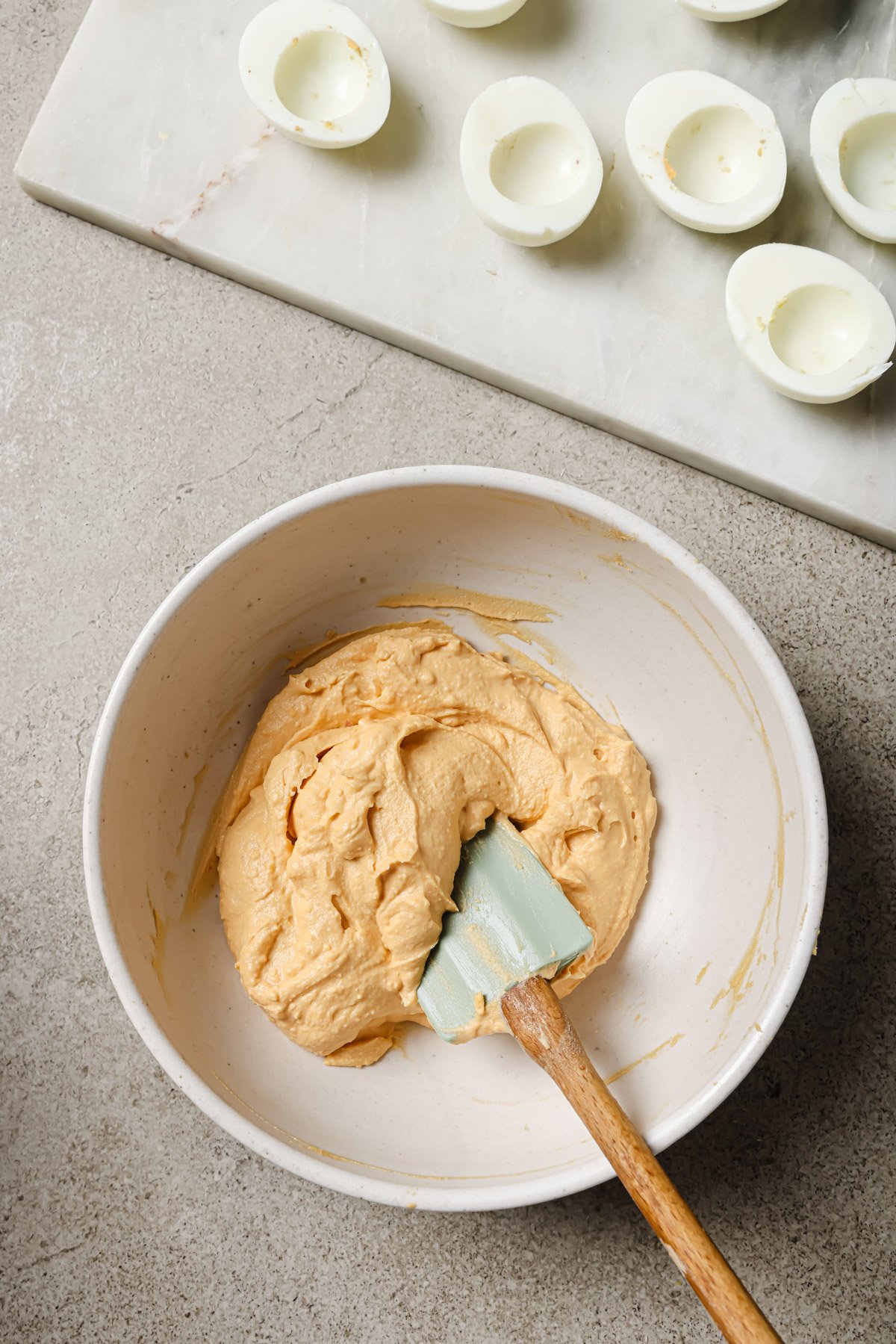 A bowl of creamy orange filling with a spatula on a countertop, beside a marble board holding several empty halved hard-boiled egg whites, prepared for making deviled eggs.