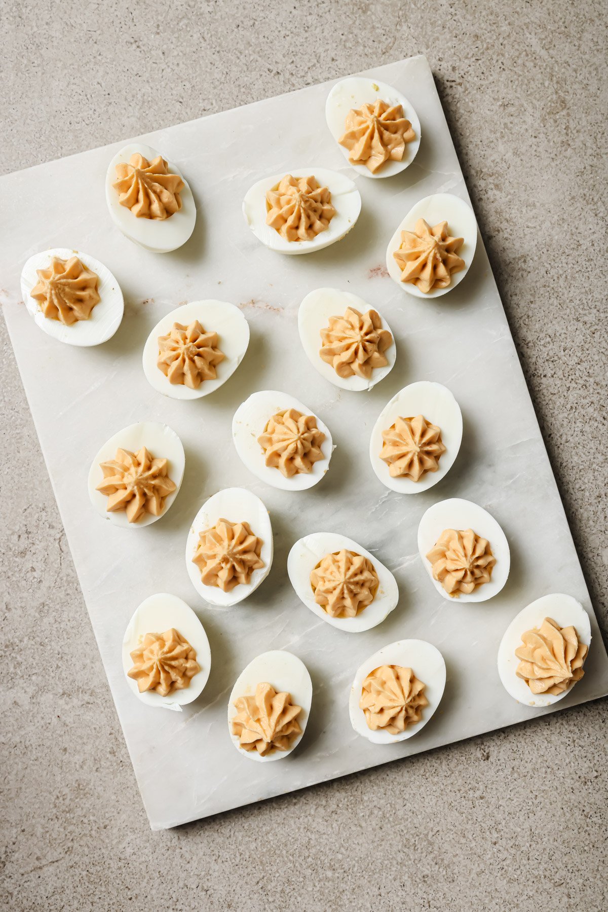A marble slab holds several deviled eggs, each topped with a neatly piped swirl of creamy filling, arranged in a loose pattern on a light-colored surface.