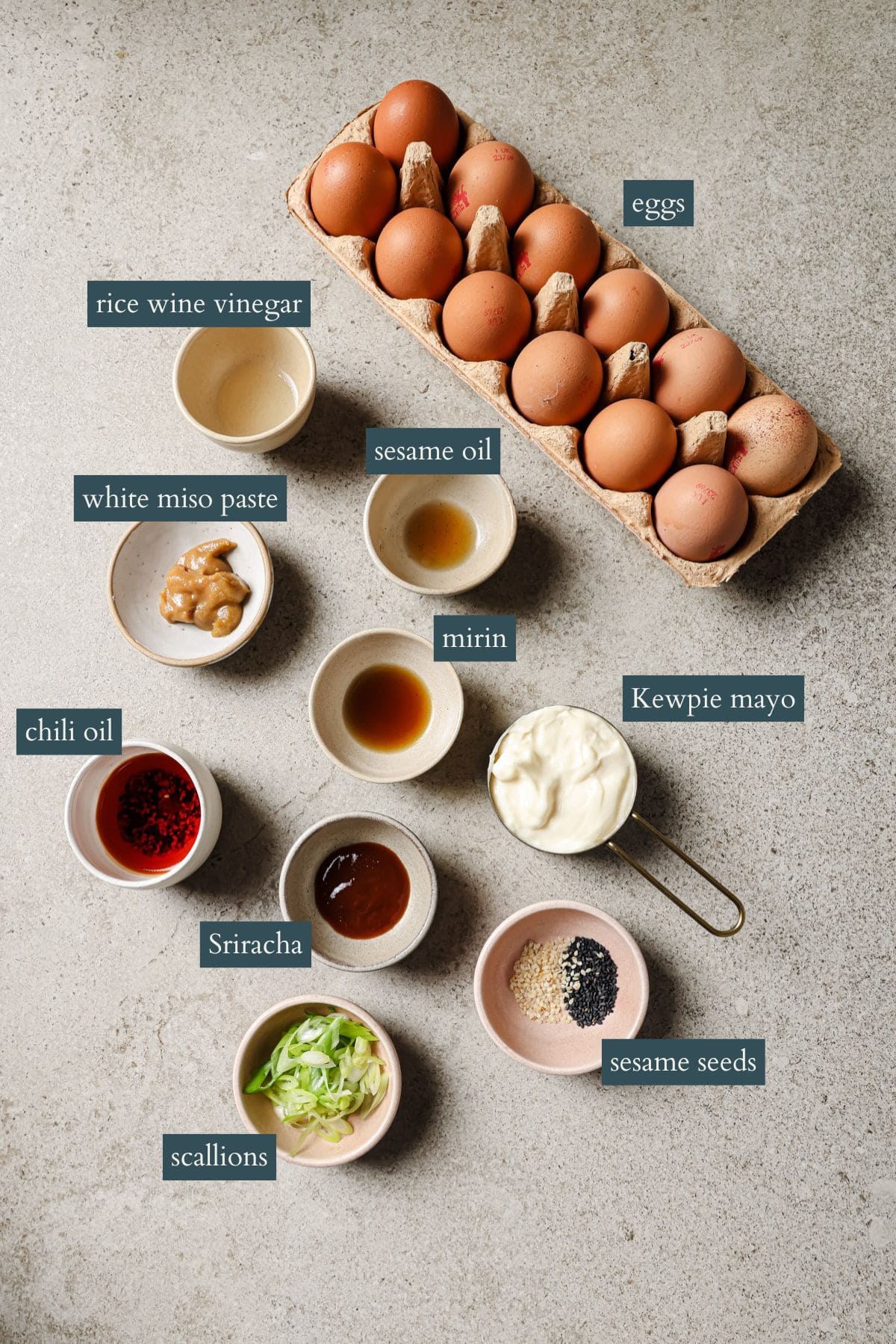 A carton of eggs sits beside small bowls containing rice wine vinegar, white miso paste, sesame oil, mirin, Kewpie mayo, chili oil, Sriracha, sliced scallions, and sesame seeds, all labeled on a gray surface.