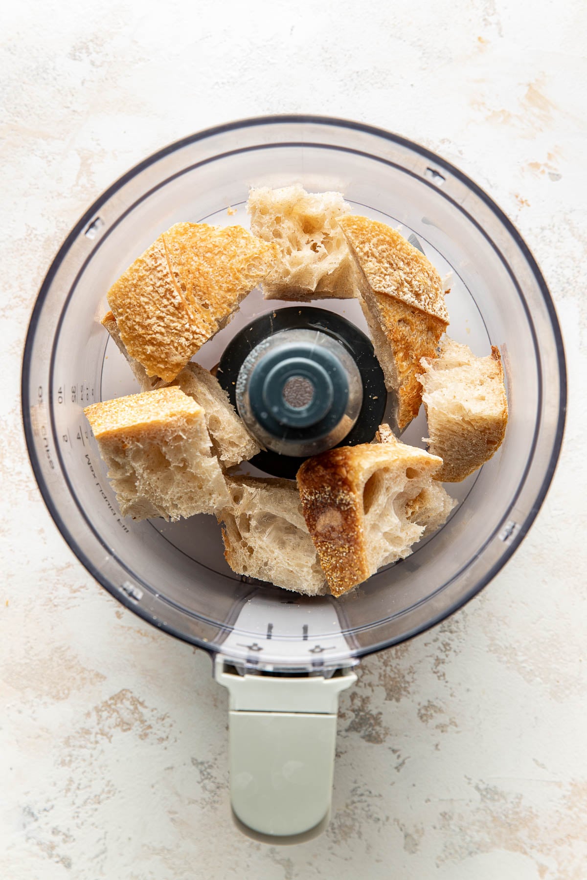 Chunks of rustic bread are arranged inside the bowl of a food processor, viewed from above on a light-colored surface.