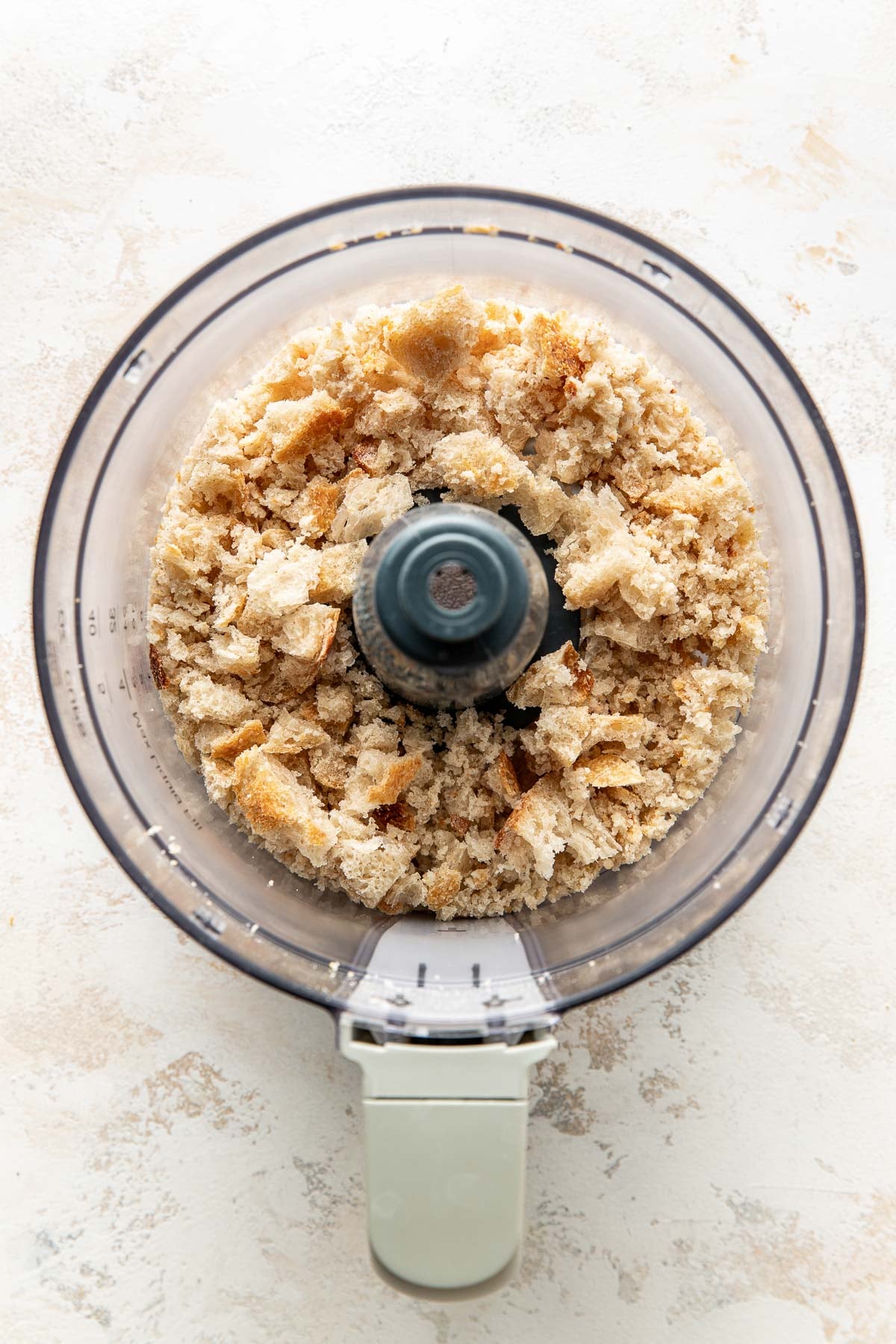 A top-down view of a food processor containing freshly made breadcrumbs from torn bread pieces on a light-colored surface.