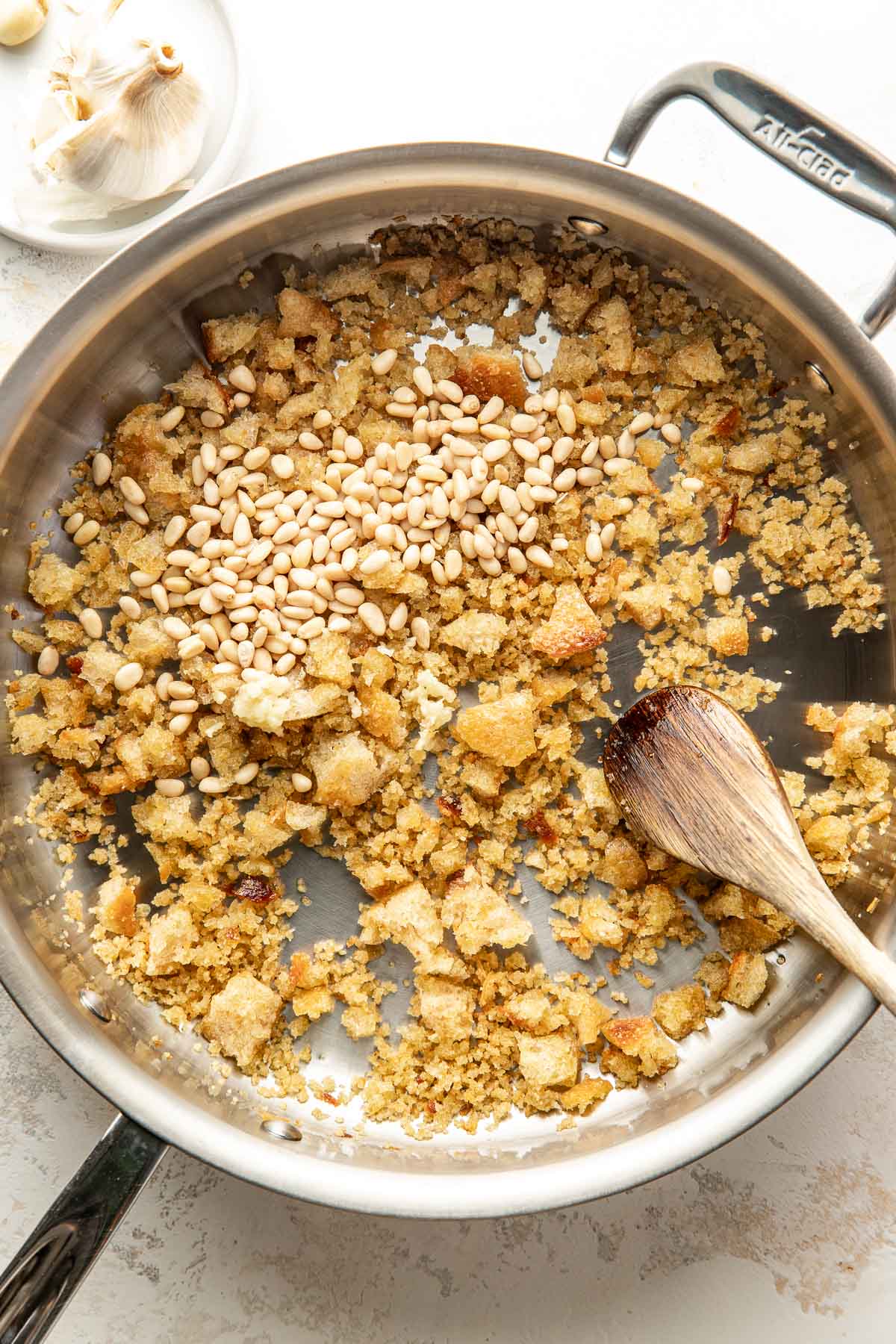 A stainless steel skillet with breadcrumb mixture and pine nuts being stirred with a wooden spoon; garlic cloves are visible in a small dish nearby.