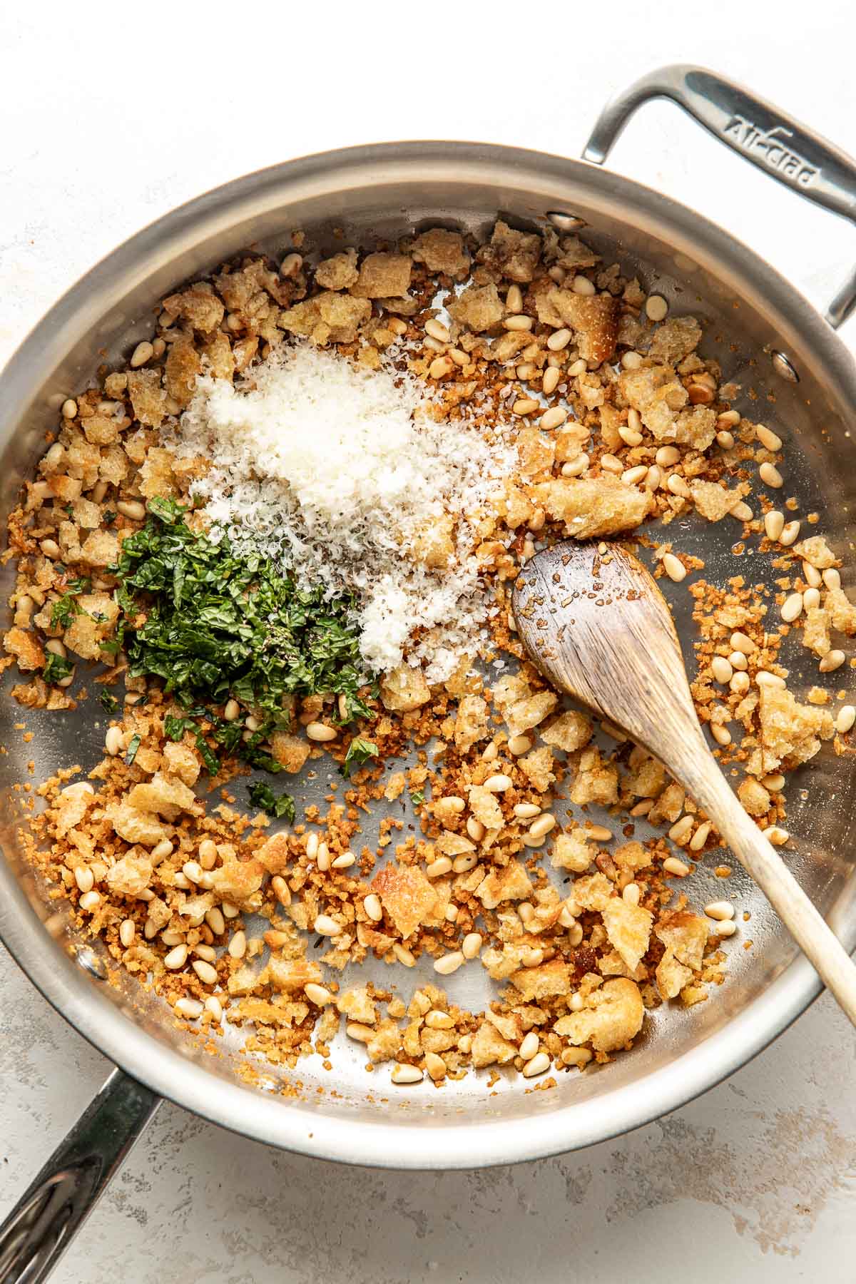 A stainless steel skillet contains toasted bread crumbs, pine nuts, chopped herbs, and grated cheese being mixed with a wooden spoon. The mixture appears to be in the process of being combined.