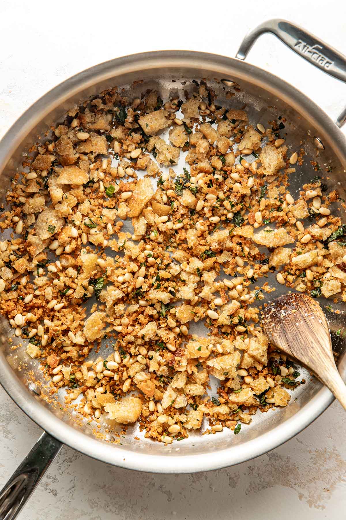 A stainless steel skillet containing toasted bread crumbs, pine nuts, and herbs being stirred with a wooden spoon.