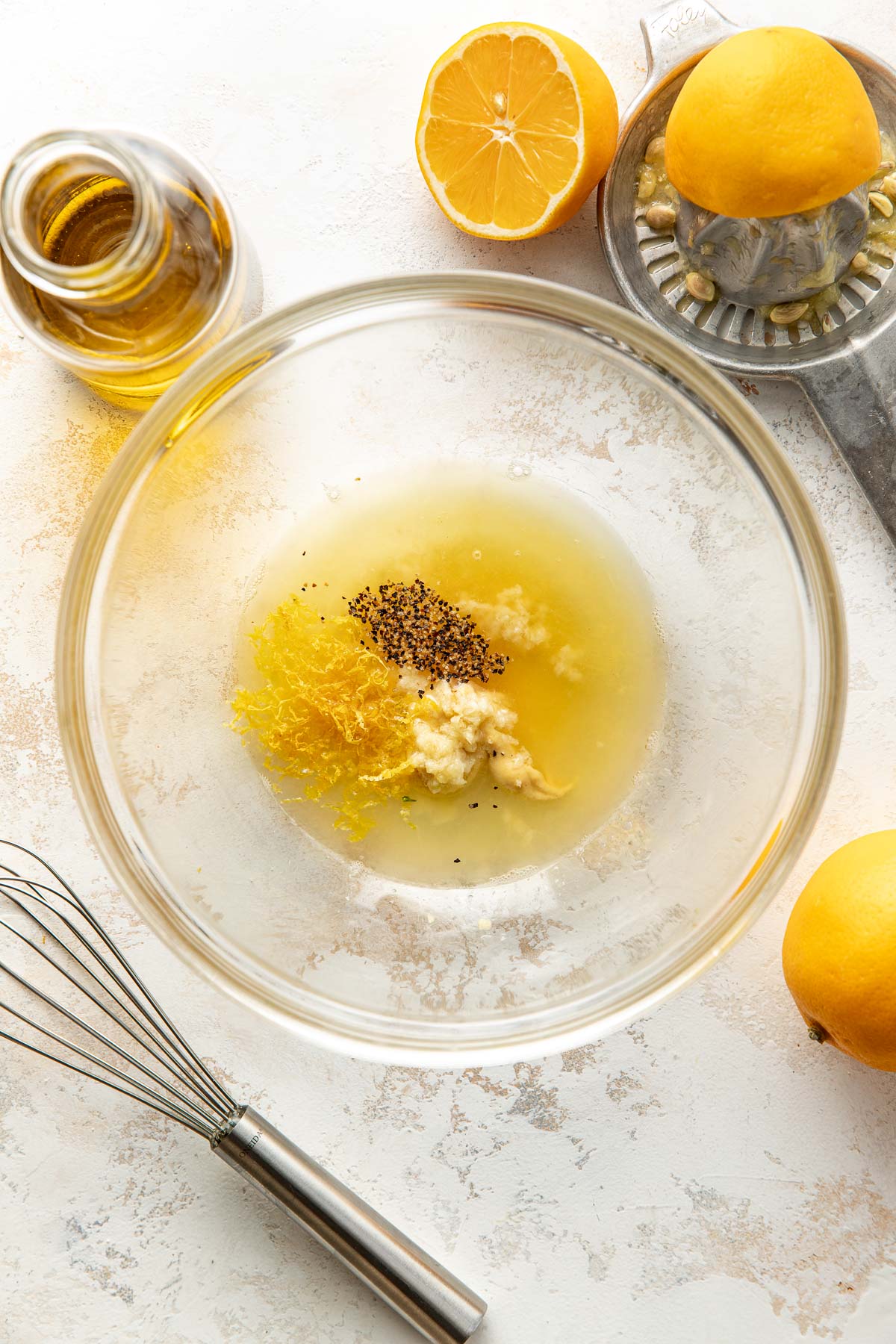 A glass bowl containing lemon zest, ground black pepper, minced garlic, and lemon juice is on a light surface. Around the bowl are a whisk, a bottle of olive oil, a halved lemon, a juicer, and whole lemons.