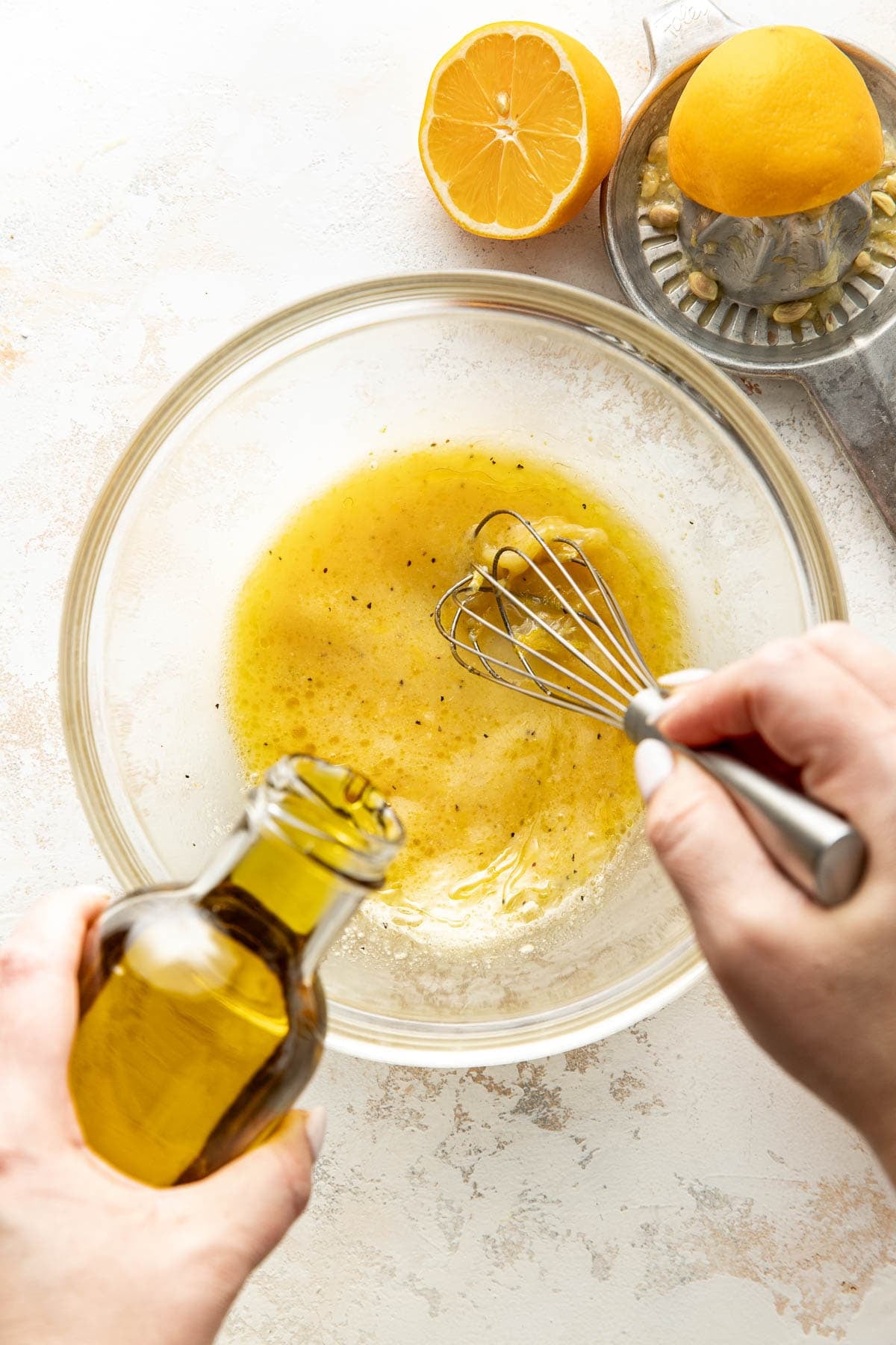 A person whisks a yellow vinaigrette in a glass bowl while pouring olive oil into it. Nearby, a halved lemon and a citrus juicer are on a light-colored countertop.