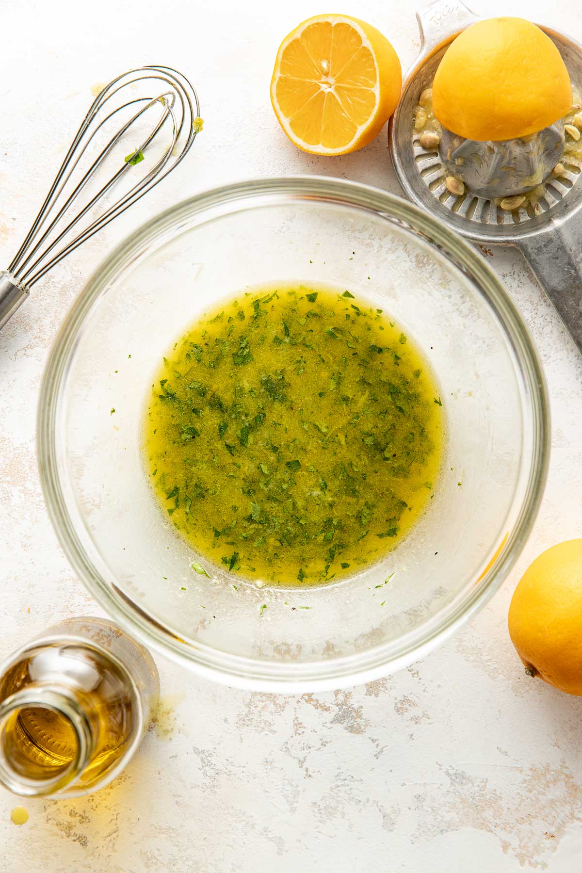 A glass bowl containing a yellow-green vinaigrette with herbs sits on a light surface, surrounded by a whisk, halved lemons, a citrus juicer, and a bottle of olive oil.