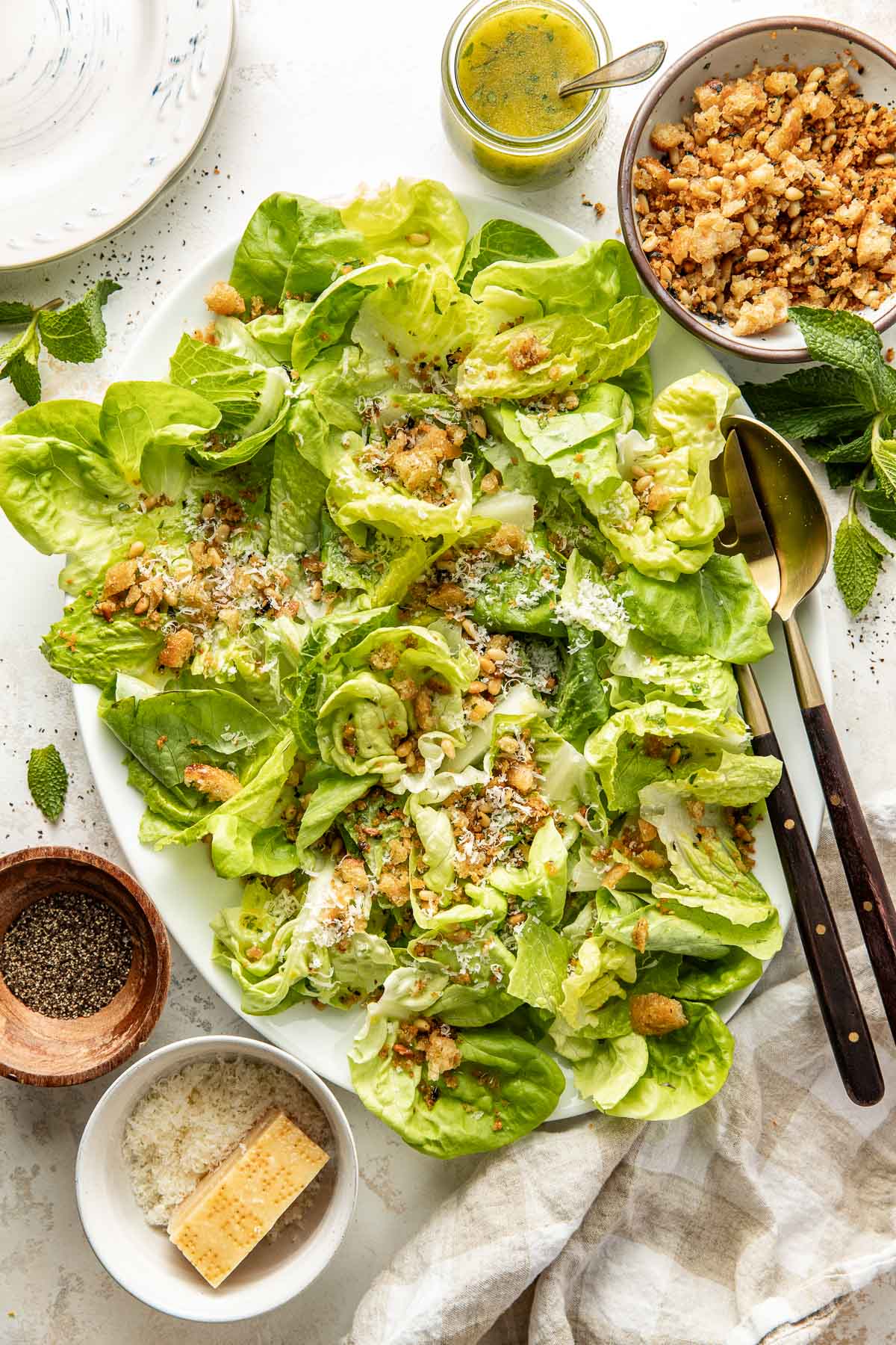 A plate of fresh green salad with lettuce, grated cheese, and croutons, surrounded by a bowl of croutons, a block of cheese, a bowl of pepper, and a jar of dressing, with utensils and a napkin on the side.