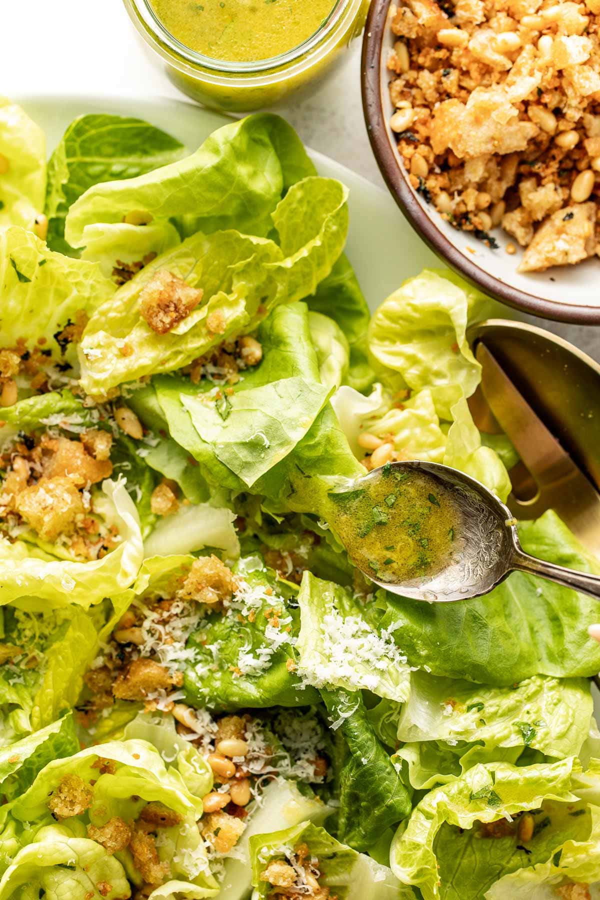 A close-up of a fresh green salad with grated cheese, toasted breadcrumbs, and dressing, next to a bowl of breadcrumbs, a jar of green salad dressing, and a spoon drizzling dressing over the salad leaves.