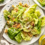 A plate of fresh green lettuce salad topped with grated cheese, croutons, and dressing. A fork rests on the plate, and other dishes and a napkin are partially visible nearby.
