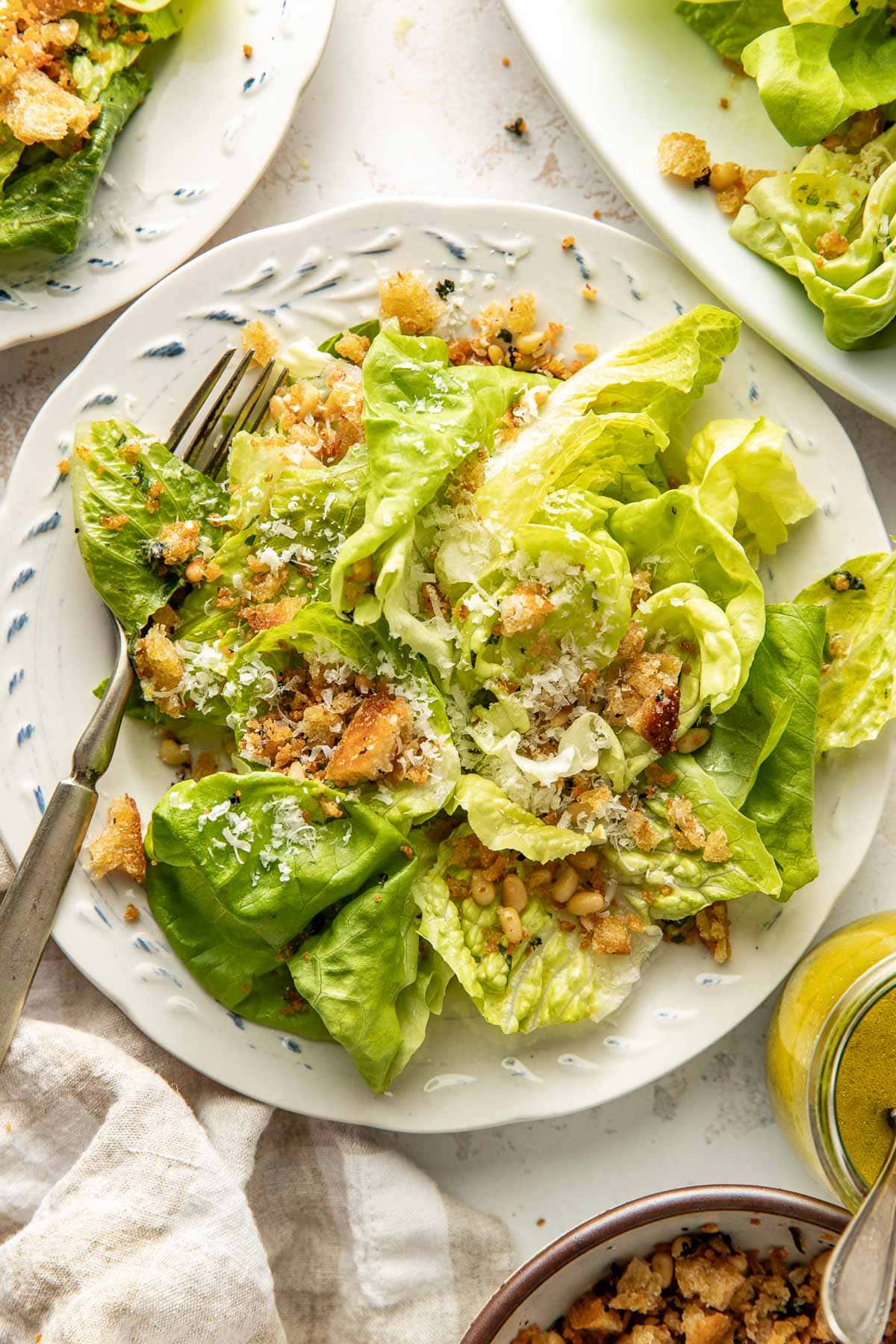 A plate of fresh green lettuce salad topped with grated cheese, croutons, and dressing. A fork rests on the plate, and other dishes and a napkin are partially visible nearby.