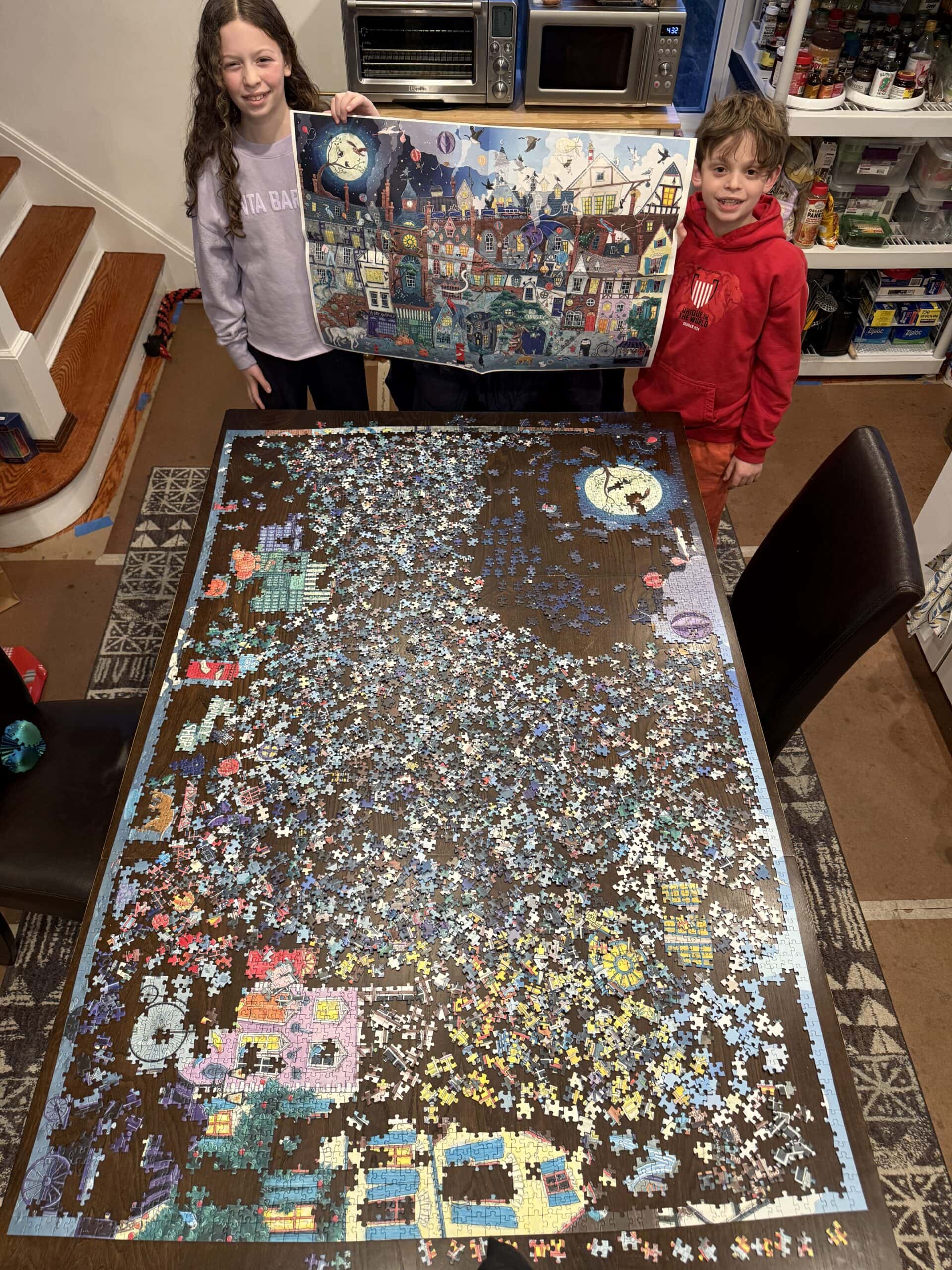 Two children stand by a table with a partially completed jigsaw puzzle. One child holds up the puzzle box showing a colorful, whimsical town scene, while many puzzle pieces are still scattered on the table.