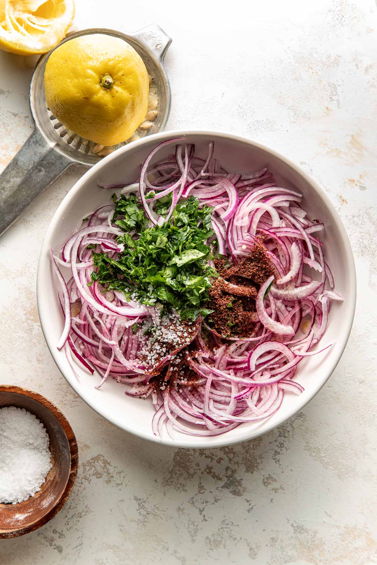 A white bowl filled with thinly sliced red onions, chopped parsley, sumac, and salt. Nearby are a halved lemon on a citrus squeezer and a small brown bowl of coarse salt.