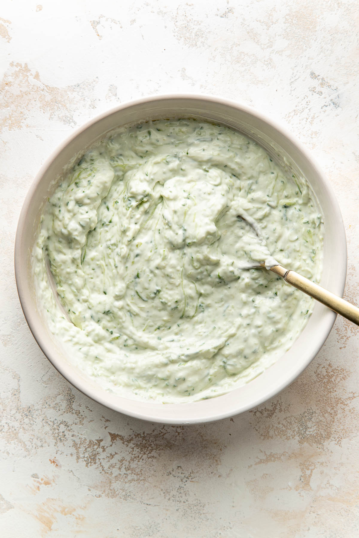 A bowl filled with creamy tzatziki sauce, speckled with green herbs, sits on a light textured surface. A spoon rests inside the bowl.