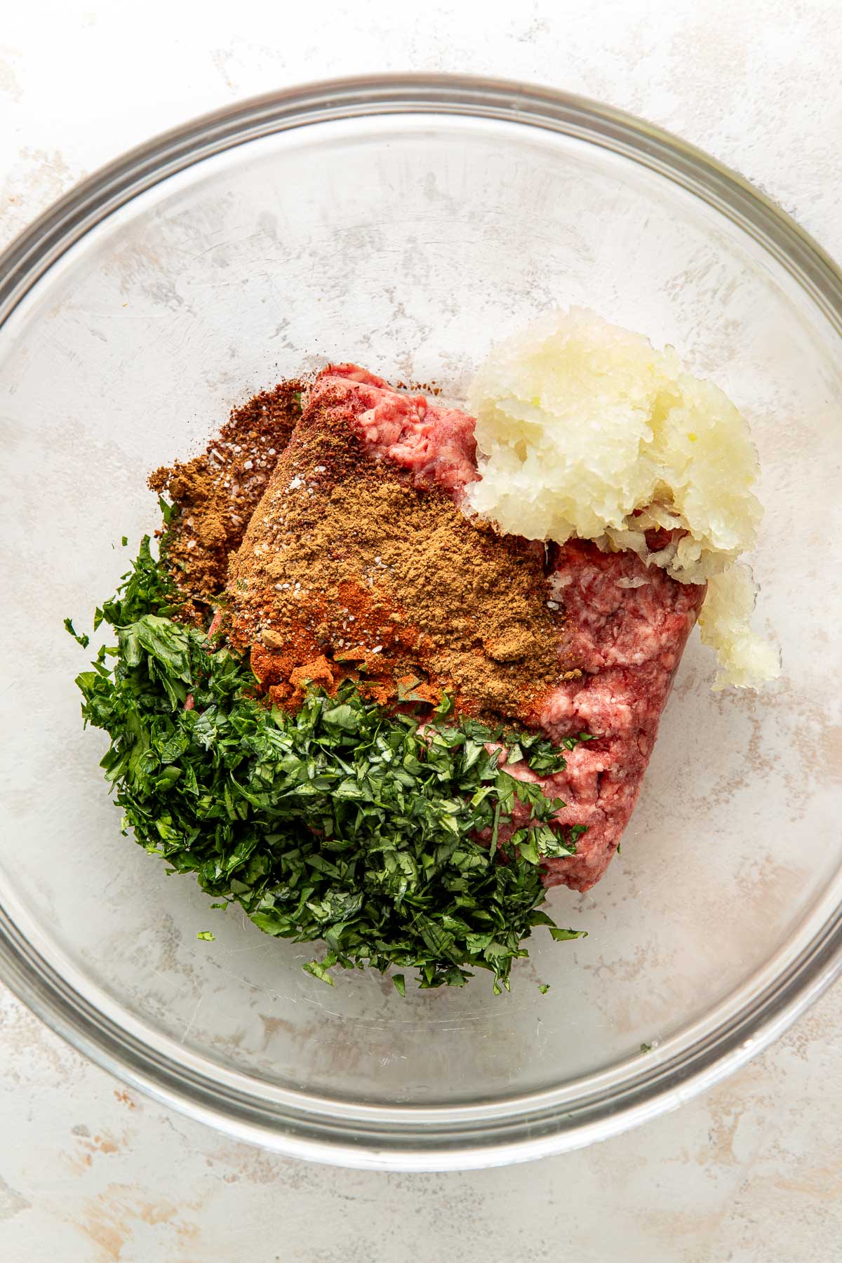 Ground meat, chopped parsley, grated onion, and a mix of spices are arranged in separate piles inside a clear glass mixing bowl, ready to be combined for a recipe.