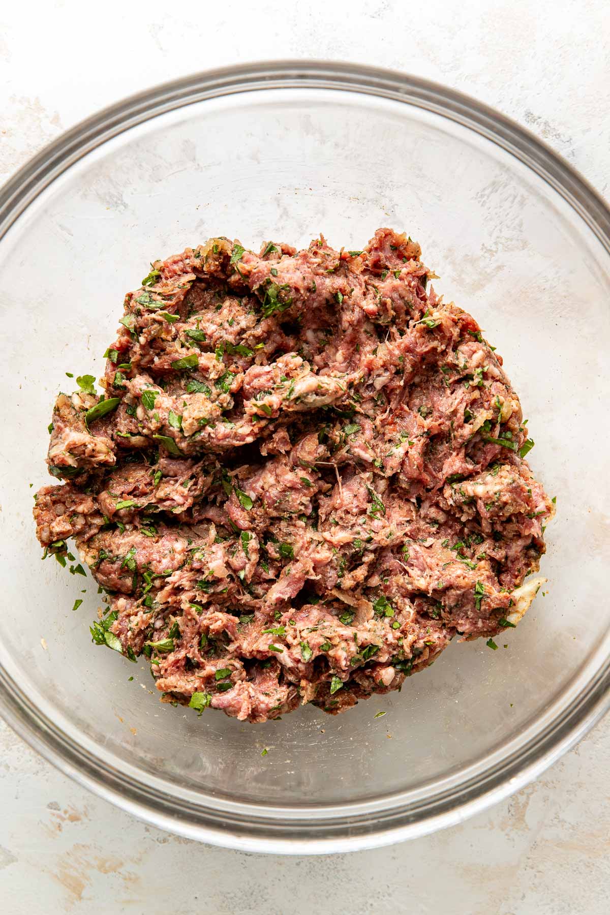 A glass bowl filled with a mixture of ground meat, chopped herbs, and seasonings, set on a light-colored surface.