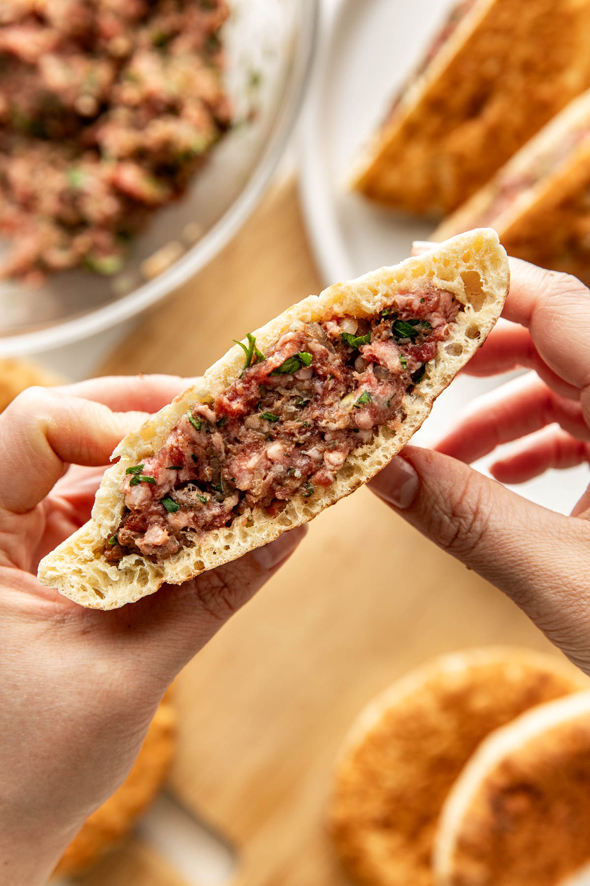 A person holds half of a pita bread filled with minced meat and herbs, showing the filling. More filled pitas and a bowl of the meat mixture are visible in the background.