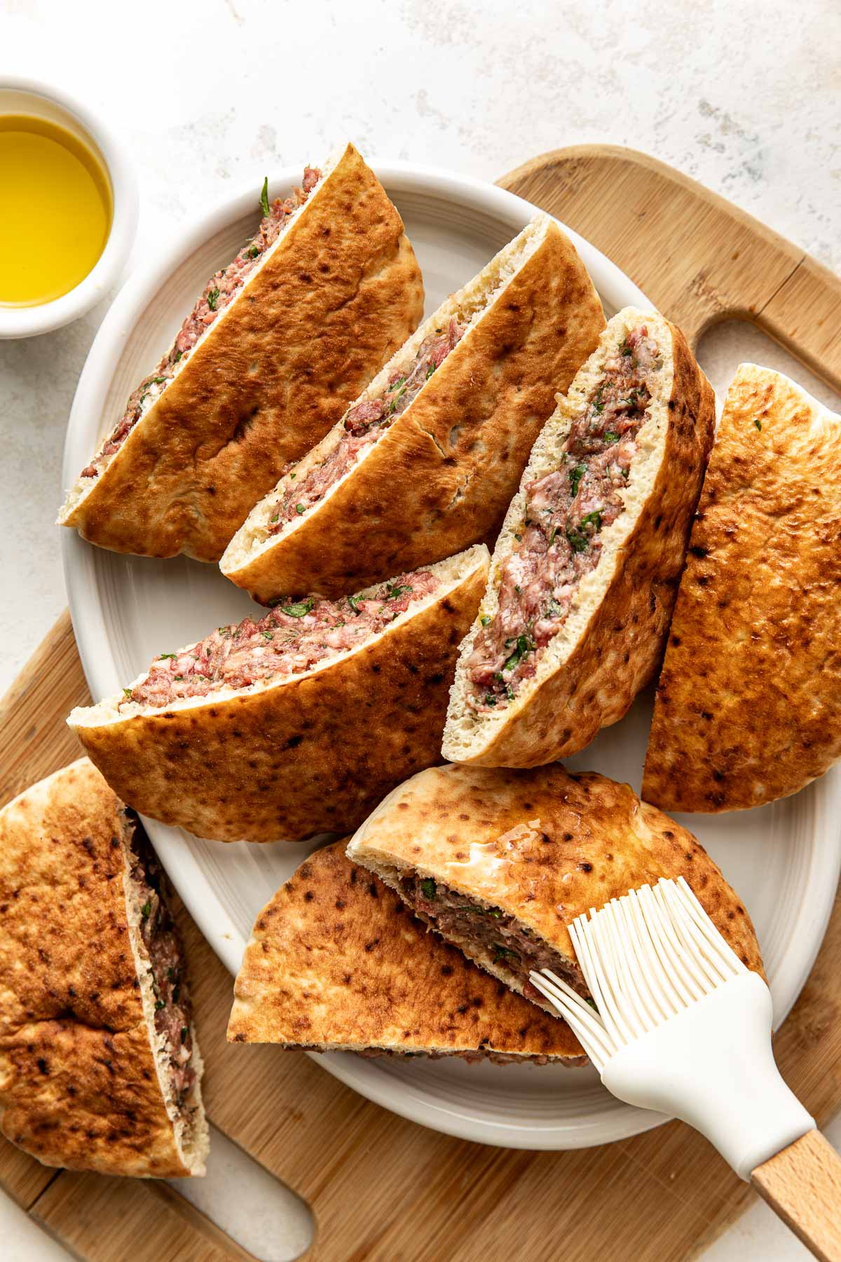 A white platter with pita bread halves filled with a seasoned meat and herb mixture. A small bowl of olive oil and a pastry brush sit nearby on a wooden board.